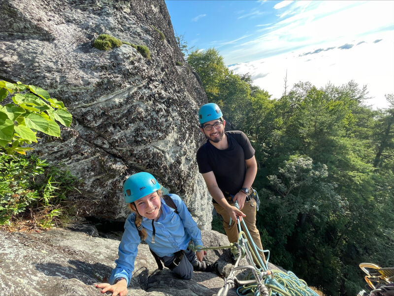 The image shows two people on a rocky cliff, likely engaged in rock climbing. The person in the foreground is wearing a blue helmet and a blue button-down shirt, kneeling on the rock. Behind them, another person stands, also wearing a helmet and a dark t-shirt. Climbing equipment is visible, suggesting they are preparing for or have just completed a climb. The background features lush green trees and a bright sky.